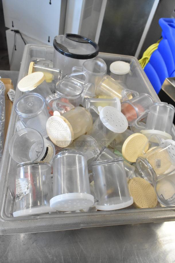 Lot of clear poly seasoning shakers with white and beige lids in a plastic bin, visible wear, mixed sizes.