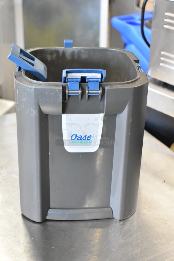 Gray Oase bin with blue locking handles, visible scratches, and "Oase Living Water" label. Good condition.