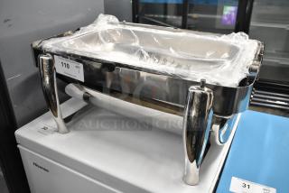 Stainless steel chafing dish with rolling lid stuck, polished handles, and plastic wrap. Auction tag visible: Item 110.