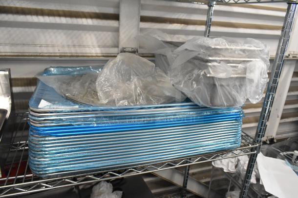 Stack of brand new metal baking pans and stainless steel drop-in bins wrapped in plastic on a metal shelf.