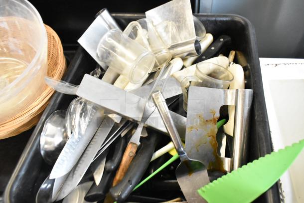 Lot of kitchen items in a black bus bin, including knives, utensils, glass mugs, and trays. Mixed condition.