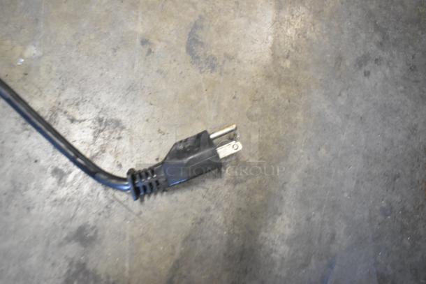 A close-up of a black two-prong electrical plug on a metal commercial countertop refrigerated beverage machine, 115 volts, 1 phase.