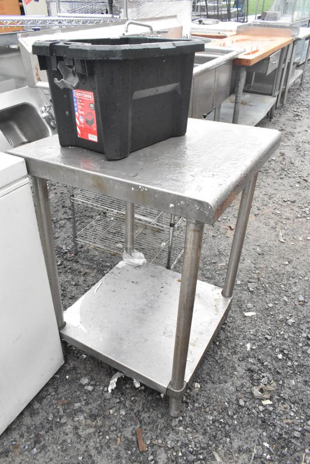 Stainless steel table with under shelf, used condition, some surface wear, located outdoors, includes black storage bin on top.