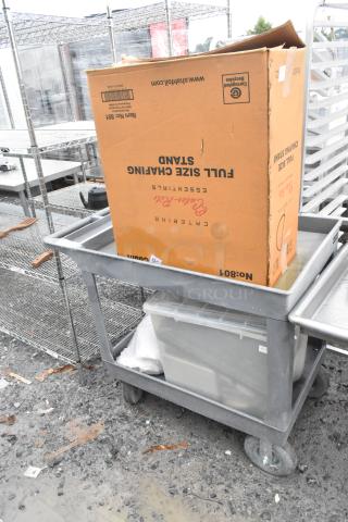 Gray poly 2-tier cart on commercial casters, sturdy design, with items stored on both shelves, outdoors on a muddy surface.