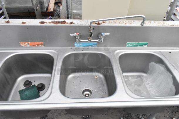 Stainless steel 3-bay sink with dual drain boards, faucet, and handles. Includes labels for washing, rinsing, sanitizing. Moderate wear.