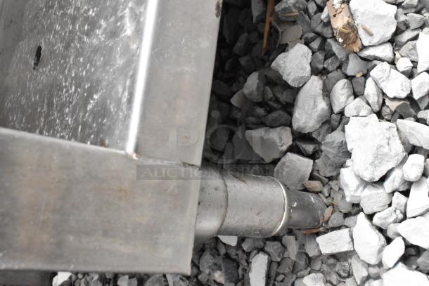 Stainless steel table corner with visible leg on gravel surface; features two metal under shelves not shown. Slight wear visible.