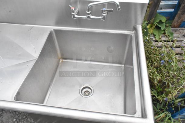 Stainless steel single bay sink with left side drain board, faucet, and handles. Slight wear, suitable for commercial use.