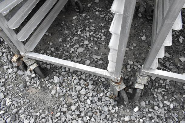 2 metal pan transport racks on commercial casters, shown on gravel. Condition: functional, some rust on casters.