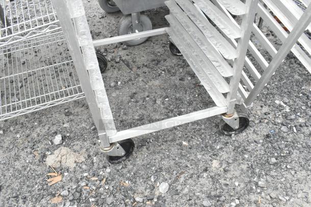 2 metal pan transport racks with commercial casters, suitable for holding trays. Appears in used condition on outdoor pavement.