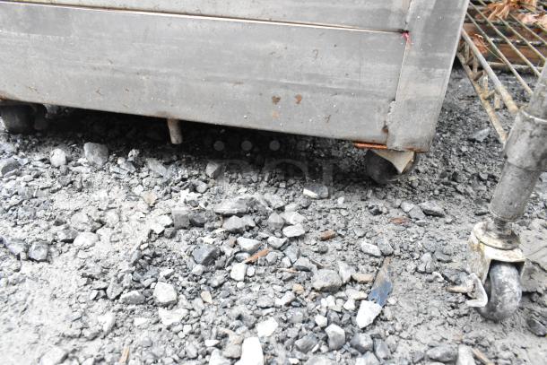 Sturdy metal rack with commercial casters on rocky ground, showing signs of rust and wear.