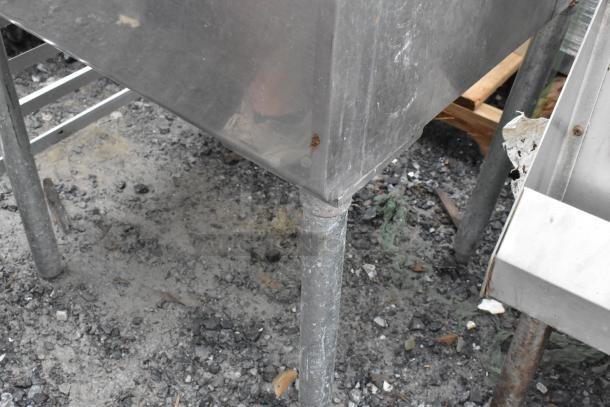 Stainless steel single bay sink with faucet and handles, showing wear and rust on leg, outdoor setting.