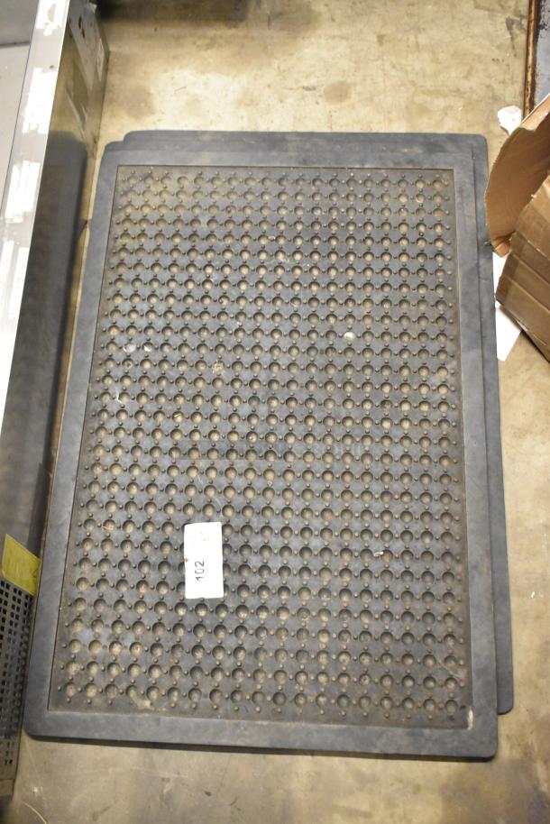 Black anti-fatigue floor mat with perforated design, visible tag labeled "102", in used condition on concrete floor.