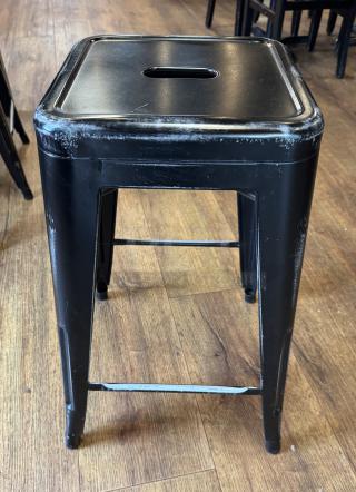 Black metal stool with distressed finish, square seat, and built-in handle, shown on wooden floor. Auction set of 4.
