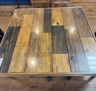 Square wooden dining table with a glass top, showcasing a multicolored wood plank design. Excellent condition, reflective glass surface.