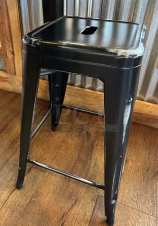 Black metal stool with a square seat and distressed finish, showing wear on edges. Suitable for industrial-style decor.