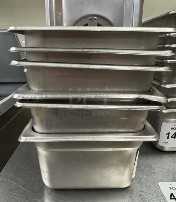 Stack of six stainless steel 1/6 pans with three lids, in good condition, showing light wear. Auction label visible.