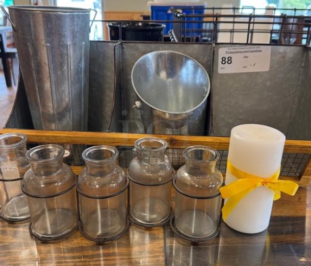 Assorted table and counter displays with metal and glass vases in various sizes, featuring a white candle with yellow ribbon.
