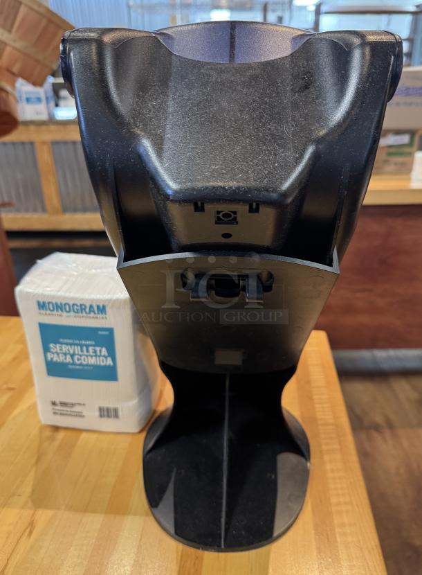 SanJamar napkin dispenser, black, includes Monogram napkins pack labeled "Servilleta Para Comida," on wooden surface.