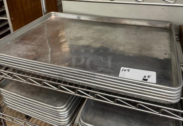 Six full-size aluminum sheet pans, used, stacked on a wire rack. Visible label indicates quantity with "x5".
