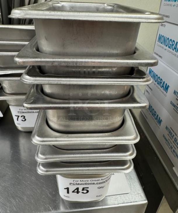 Seven stainless steel pans, 1/6 x 4 size, stacked, auction tag "145" visible, used condition.