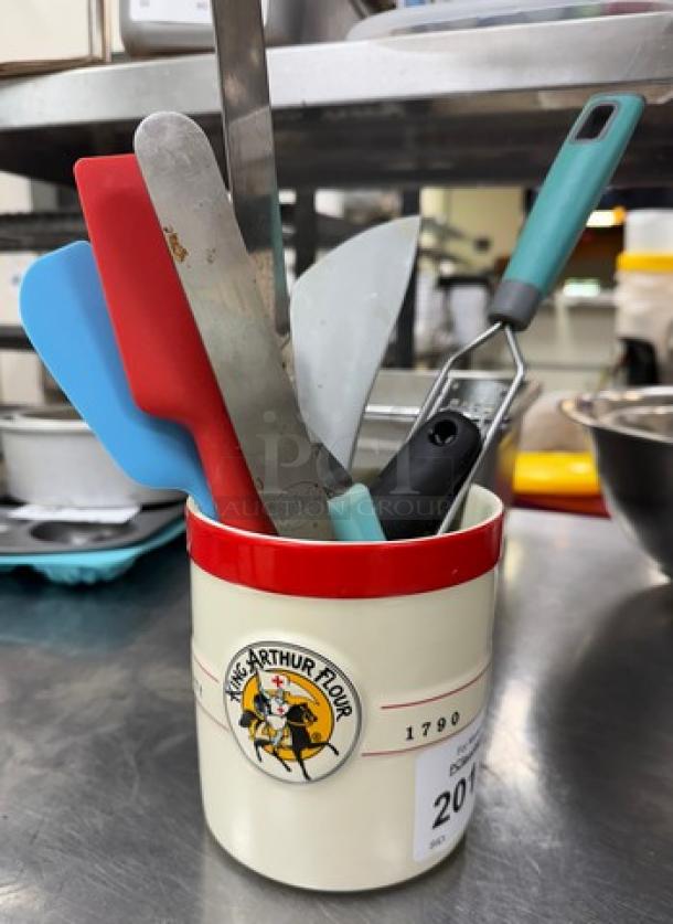 Assorted kitchen tools in a King Arthur Flour container, features spatulas and a peeler; tools in used condition.