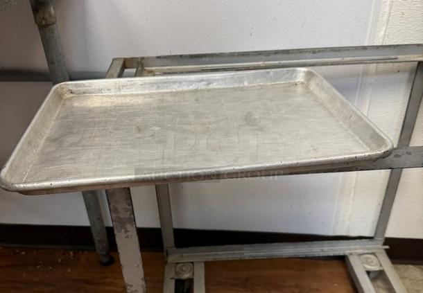 Six half-size sheet pans in used condition, showing signs of wear. Made of aluminum, stored on metal rack.