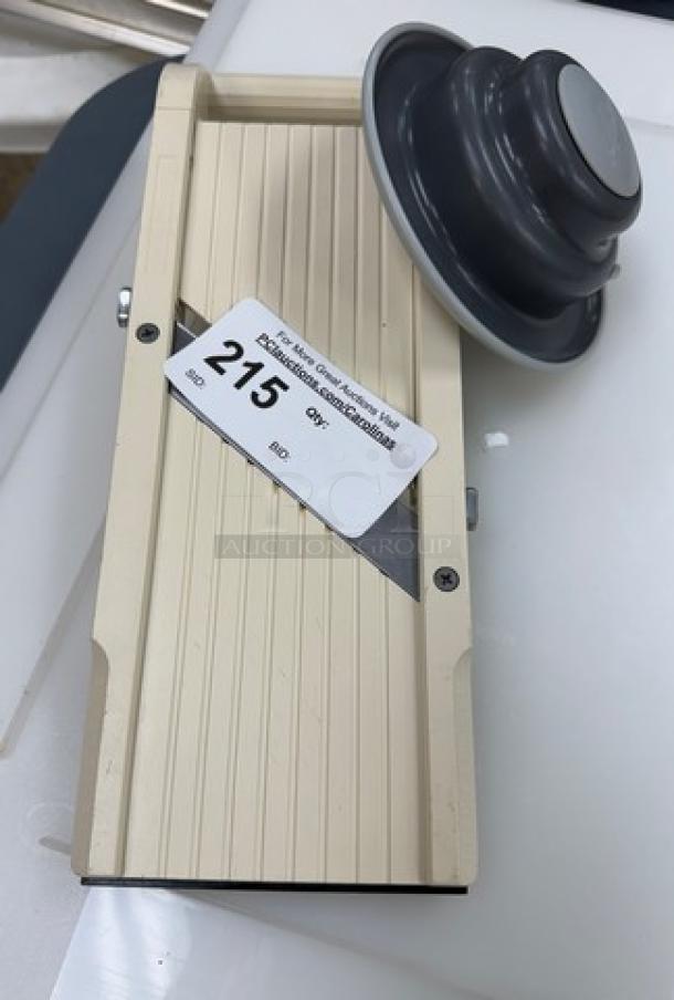 Mandolin slicer with beige body and black food holder. Slightly used, shows auction label number 215. Durable kitchen tool.