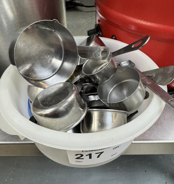 Assorted metal measuring cups and spoons in a white container, labeled "217", in used condition.