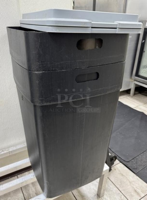 Large black trash can with gray lid, rectangular shape, used condition with visible scratches, suitable for outdoor or commercial use.