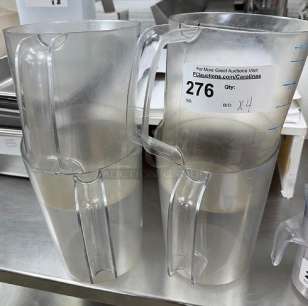 Four clear plastic 1-gallon pitchers stacked. Each shows measuring lines and has a large handle. Bid tag indicates auction lot 276, quantity 4.