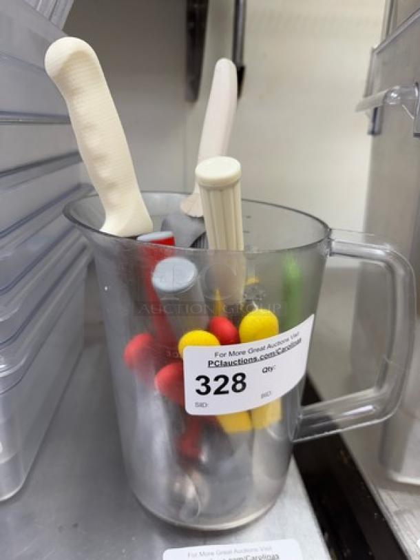 Clear pitcher with assorted scoops, spreaders, and bread knives, various colors, auction tag labeled "328."