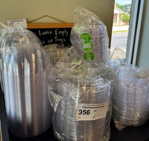 New 32 oz clear plastic bowls with lids, wrapped in plastic. Includes multiple stacks, labeled for auction, item number 956.