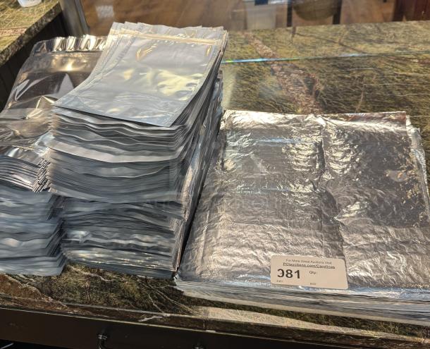 Stacked foil sealable bags, shiny silver, large quantity. Auction label "381" visible. Ideal for storage and preservation.