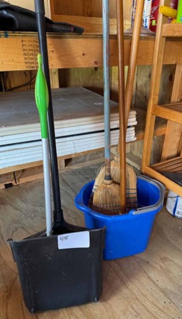 Assorted cleaning supplies including a broom, mop, and dustpan in a blue bucket labeled "4/15" on the dustpan.
