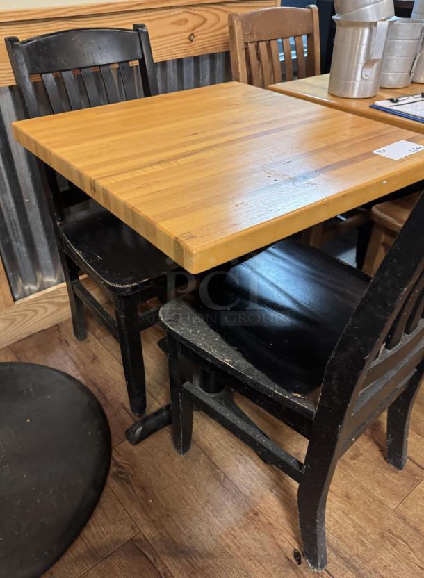 Wooden dining table with two black chairs. Table surface shows moderate wear. Chairs feature a classic slatted back design.