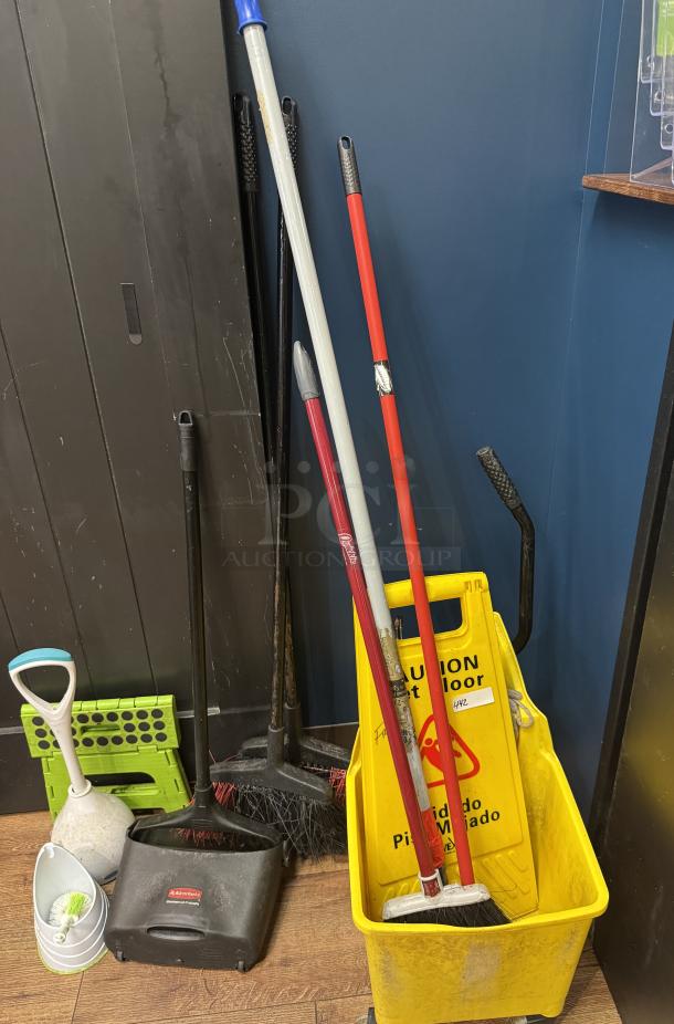 Cleaning supplies including mops, brooms, yellow caution sign, dustpan, brushes, and bucket. Items show signs of use.