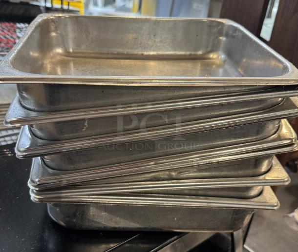 Nine stainless steel half-size sheet pans stacked, showing slight wear. Durable construction, ideal for baking or roasting.