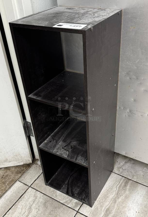 Black bookcase with 3 shelves, showing slight wear and dust. Compact design, suitable for small spaces. Label visible on top.