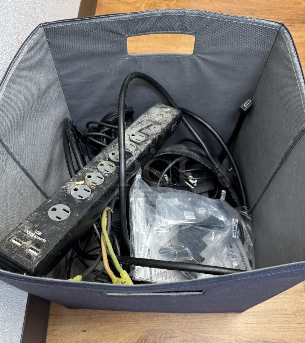 A box of assorted cables, cords, and chargers, including a power strip with USB ports and various electrical connectors.