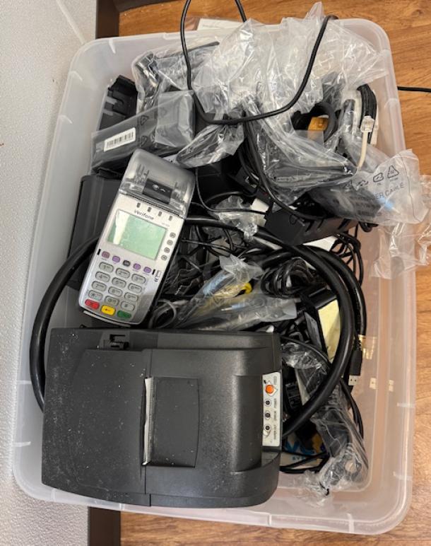 Assorted electronics including cables, terminals, printers, and chargers in a plastic bin. Some items are in original packaging.