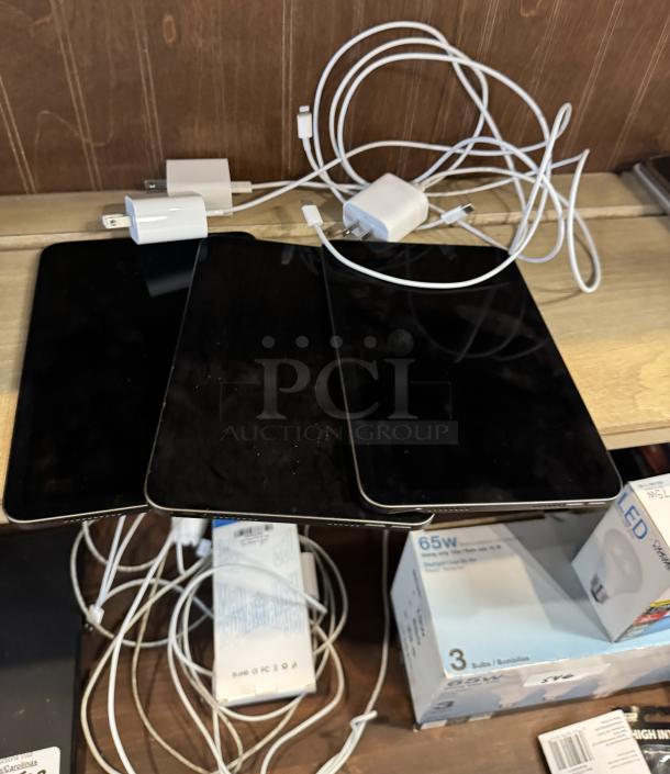 Three Apple iPads Mini with chargers on wooden surface, tested and working condition. Includes three white charging cables.