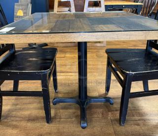 Wooden dining table with glass top, features dark wood grain and metal pedestal base. Accompanied by two worn black chairs.