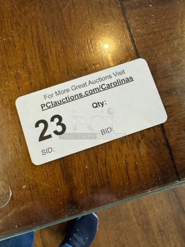 Wooden dining table with a glass top, showing natural wood grain. Auction tag: "23," URL: "PClauctions.com/Carolinas."