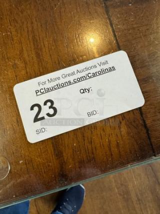 Wooden dining table with a glass top, showing natural wood grain. Auction tag: "23," URL: "PClauctions.com/Carolinas."