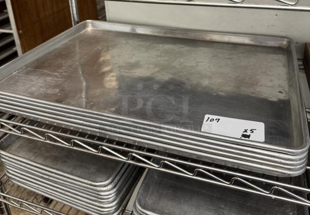 Full-size sheet pans, lot of 6, stacked on metal rack. Visible label marked "107 x5". Used condition with surface wear.