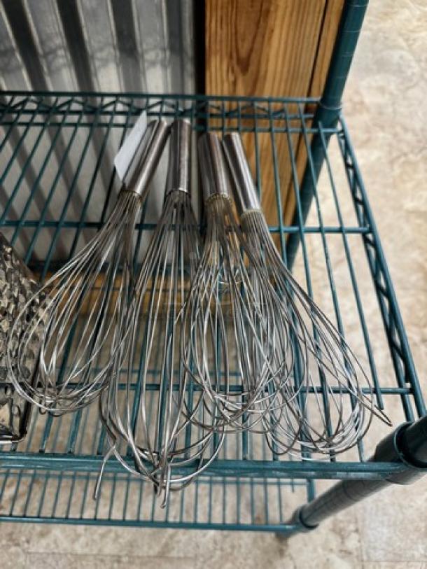 Set of five large stainless steel whisks, metal handles, excellent condition, displayed on a wire shelf.