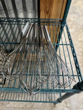 Set of five large stainless steel whisks, metal handles, excellent condition, displayed on a wire shelf.