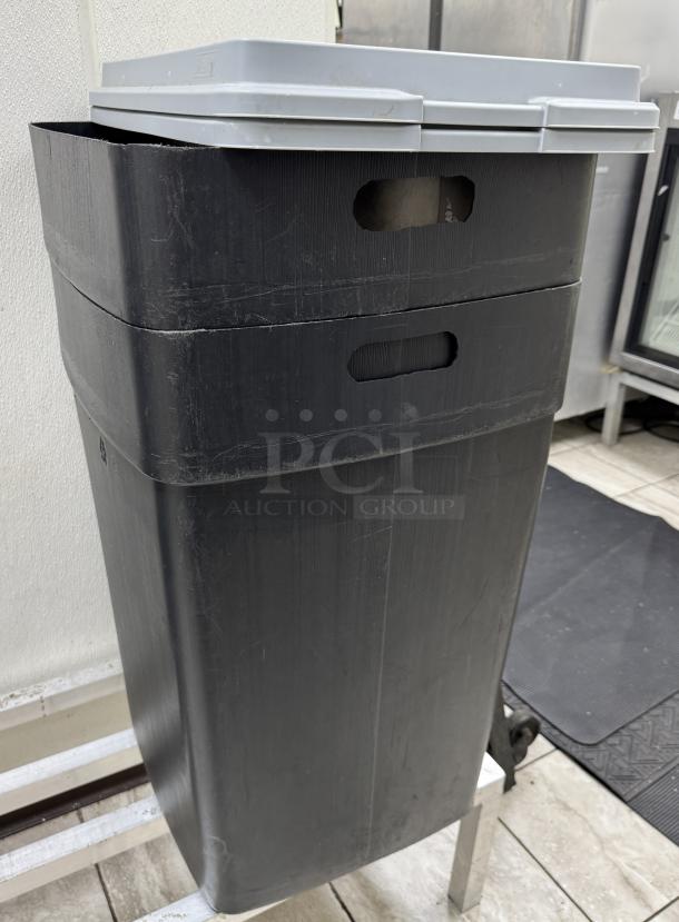 Large black trash can with a grey lid, showing signs of wear. Features integrated handles for easy handling. Auction lot of 4.