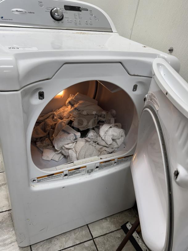 White Whirlpool Cabrio clothes dryer, open door, interior light on, towels inside. Features control panel, some surface dirt.