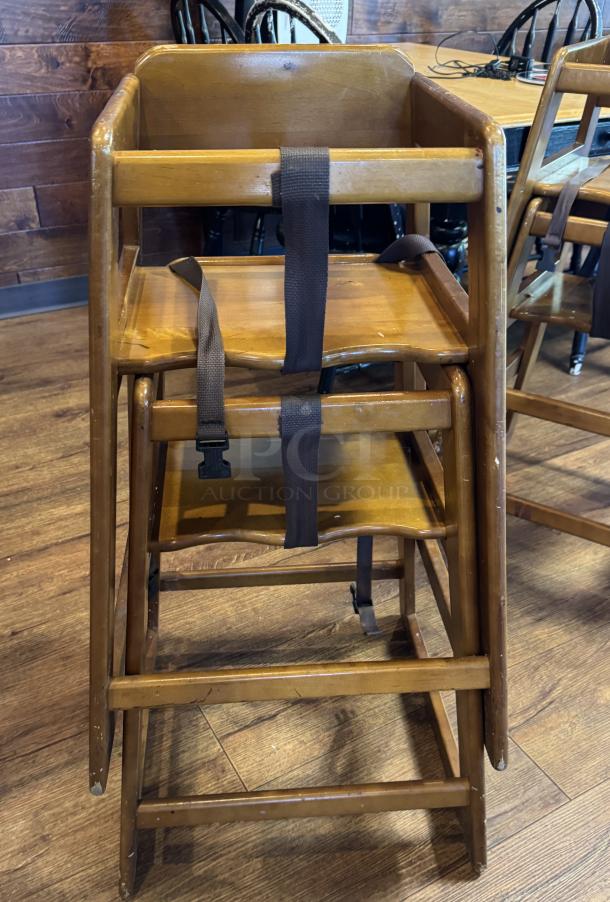 Two wooden high chairs, stacked, with safety straps and visible wear marks. Sturdy design, ideal for restaurant use.